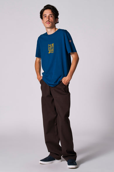 Blauw T-shirt met korte mouwen van Cyclo Club Marcel, met gele print en gedragen met bruine broek en blauwe sneakers.
