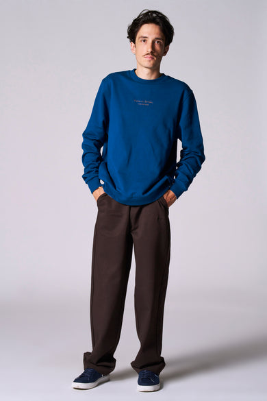 Pull-over bleu avec col rond de Cyclo Club Marcel, porté avec un pantalon marron et des baskets bleues.