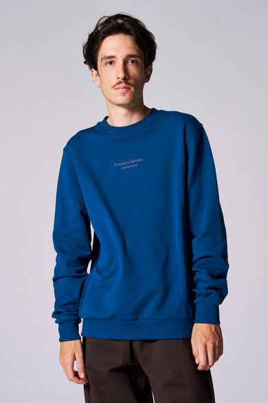 Pull-over bleu avec col rond de Cyclo Club Marcel, imprimé texte sur la poitrine, associé à un pantalon marron.
