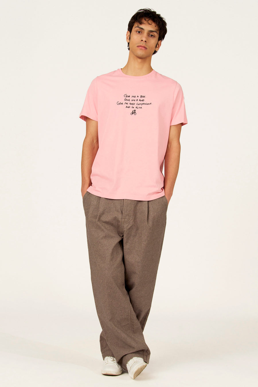 T-shirt à manches courtes rose de Cyclo Club Marcel, avec impression de texte noir et dessin de vélo, porté avec un pantalon marron.