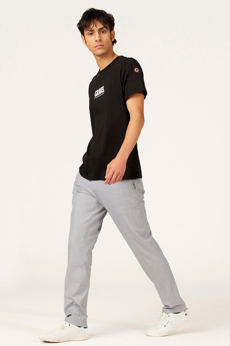 T-shirt à manches courtes noir de Cyclo Club Marcel, avec imprimé blanc et porté avec un pantalon gris et des baskets blanches.