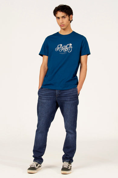 T-shirt bleu à manches courtes de Cyclo Club Marcel, avec imprimé blanc, combiné avec un jean foncé et des baskets.