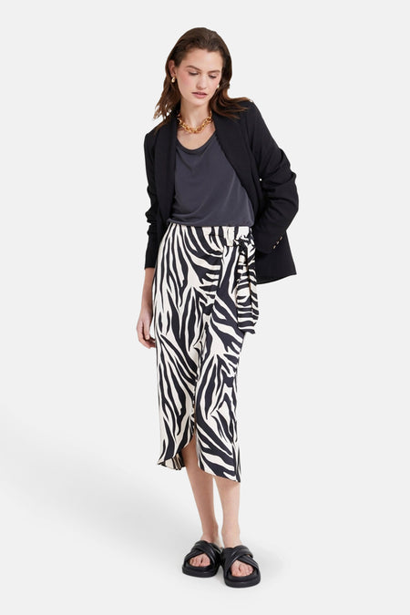 Lange multicolor rok van NAF NAF, met zebraprint en een knoopdetail, gecombineerd met een zwarte blazer en sandalen.
