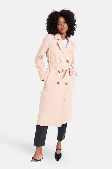 Beige lange jas van NAF NAF, met een strikceintuur, gecombineerd met een zwarte broek en hakken.
