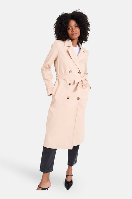 Beige lange jas van NAF NAF, met een strikceintuur, gecombineerd met een zwarte broek en hakken.
