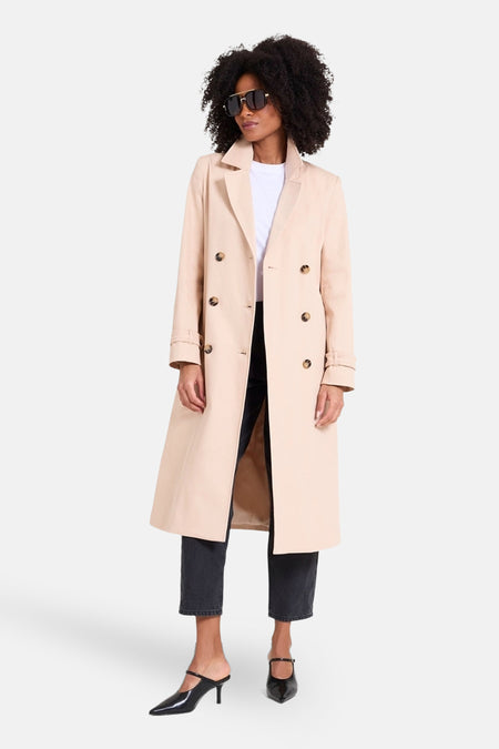 Beige lange jas van NAF NAF, met dubbele knopenrij, gedragen met een wit shirt en een zwarte jeans.
