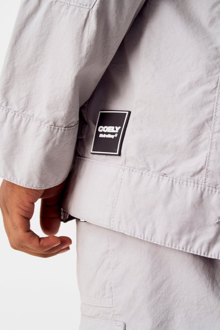 Grijze jas van COELY, met een vierkant logo op de borst en details van stiksels.