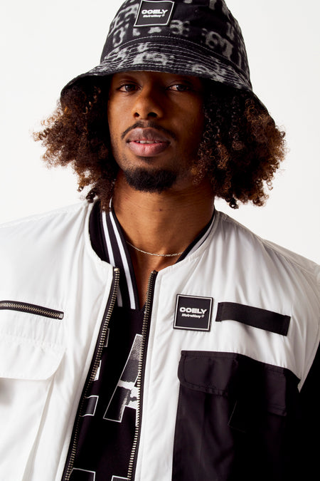 Manteau blanc de COELY avec des accents noirs et une fermeture éclair remarquable, porté avec un chapeau bucket imprimé et une chaîne d'argent.