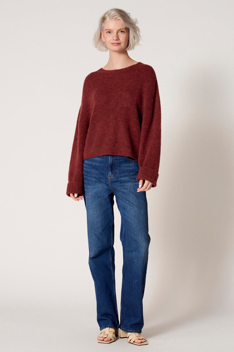 Bordeaux trui met ronde hals van Clouds of Fashion, gedragen met een blauwe jeans en beige sandalen.
