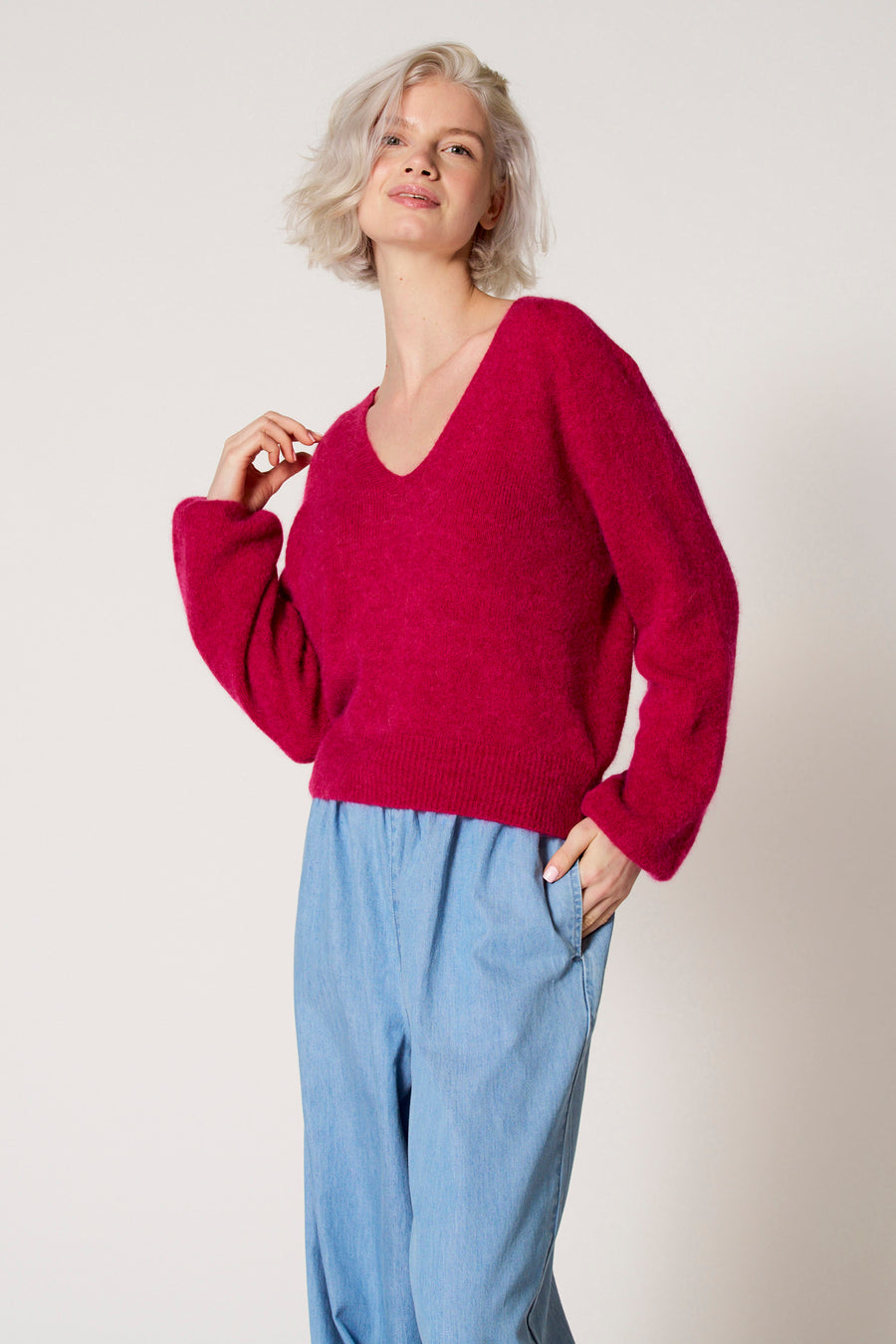 Roze trui met V-hals van Clouds of Fashion, gedragen met lichtblauwe broek en een hand in de zak.
