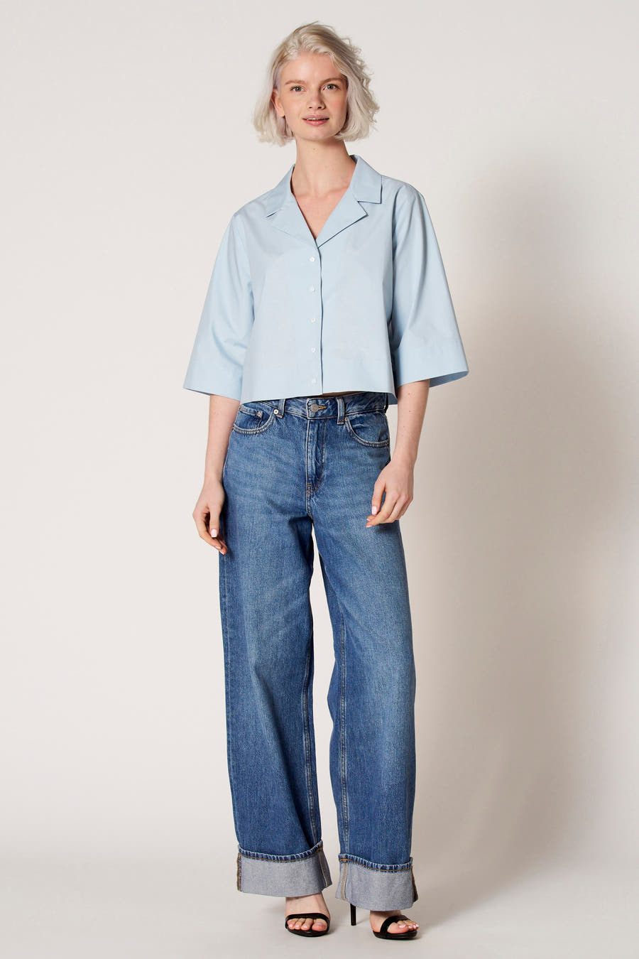 Blouse bleue à manches trois-quarts de Clouds of Fashion, avec col ouvert, portée avec des jeans larges et des baskets.