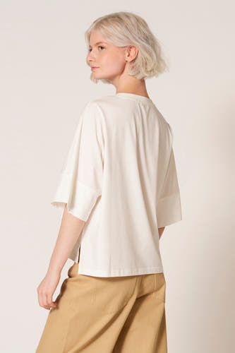 Ecru T-shirt met korte mouwen van Clouds of Fashion, met wijde mouwen en een subtiele split aan de zijkant, gecombineerd met een bruine broek.
