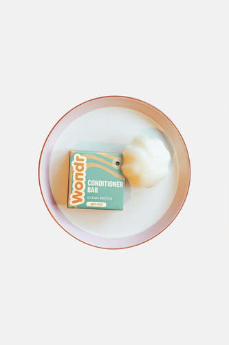 Witte schelpvormige conditioner bar van WONDR CARE, naast de teal verpakking met oranje golven, op een wit dienblad met roze rand.