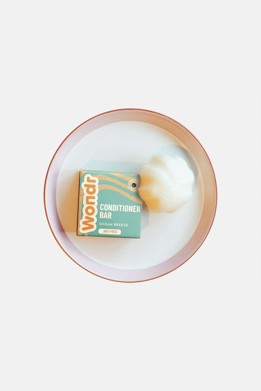 Witte schelpvormige conditioner bar van WONDR CARE, naast de teal verpakking met oranje golven, op een wit dienblad met roze rand.