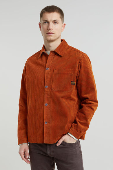 Een man in een oranje corduroy overhemd met bruine broek tegen een lichte achtergrond.