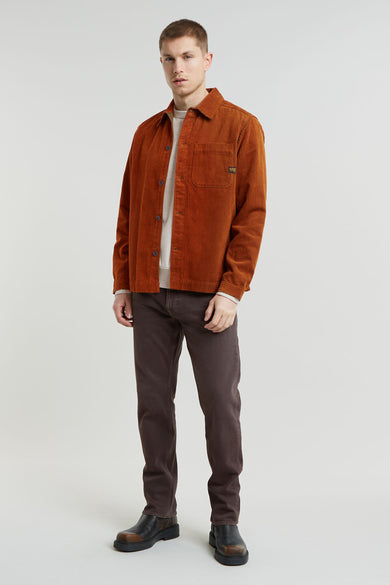 Man in oranje ribfluwelen overhemd, bruine broek en leren laarzen staand tegen een witte achtergrond.