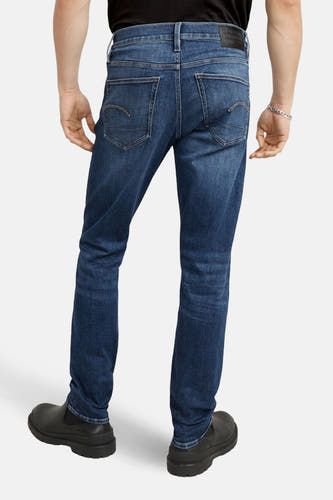 Slim jeans van blauw denim van G-Star RAW, achterkant gezien, met donkere schoenen en zilveren armband.