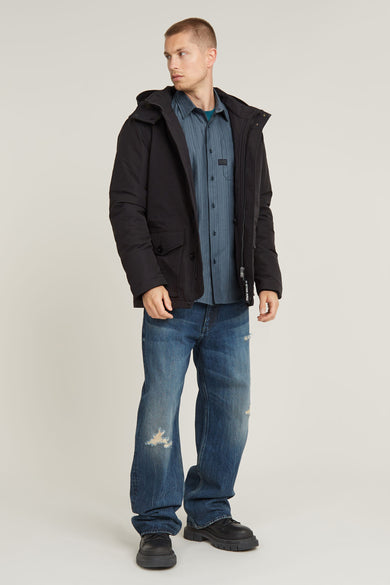 Zwarte lange jas van G-Star RAW, gedragen over een blauw gestreept overhemd en losse jeans met gaten.
