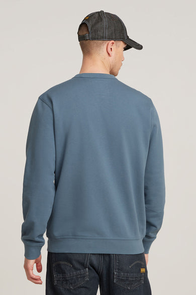 Blauwe sweater met ronde hals van G-Star RAW, gezien van achteren, met donkere jeans en een denim pet.
