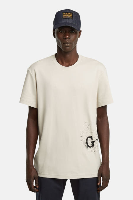 Ecru T-shirt met korte mouwen van G-Star RAW, met een print aan de onderkant en gedragen met een donkerblauwe pet.