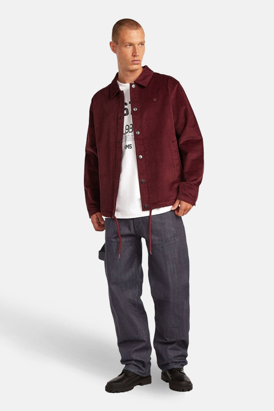 Model draagt bordeauxrood corduroy jasje, wit T-shirt en wijde blauwe jeans met zwarte schoenen.