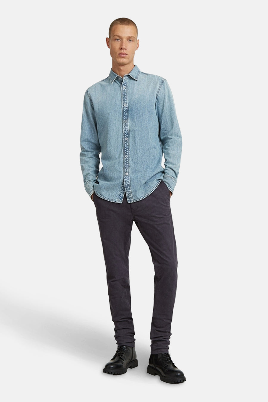 Man, staand, denim overhemd, donkere broek, zwarte schoenen.