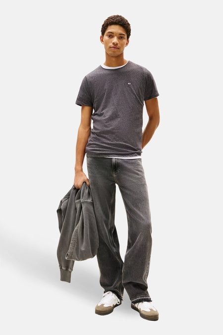 Man in grijze jeans en T-shirt met jas in de hand, staand op witte achtergrond.