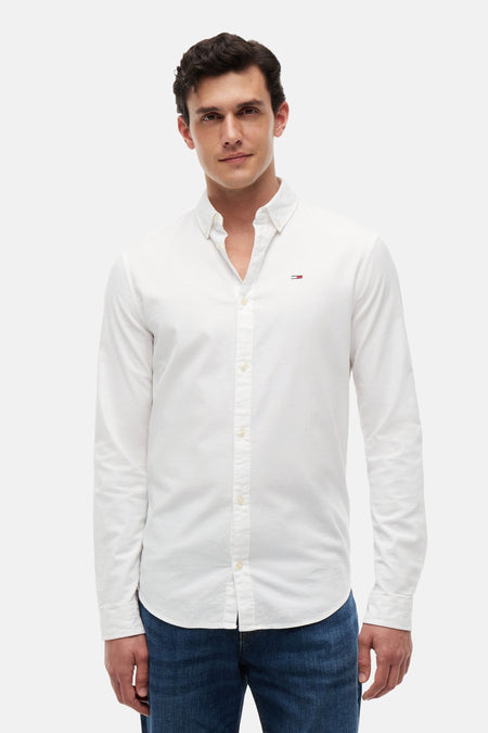 Chemise blanche à manches longues TOMMY JEANS, avec petit logo drapeau sur la poitrine, col boutonné, portée avec un jean bleu.