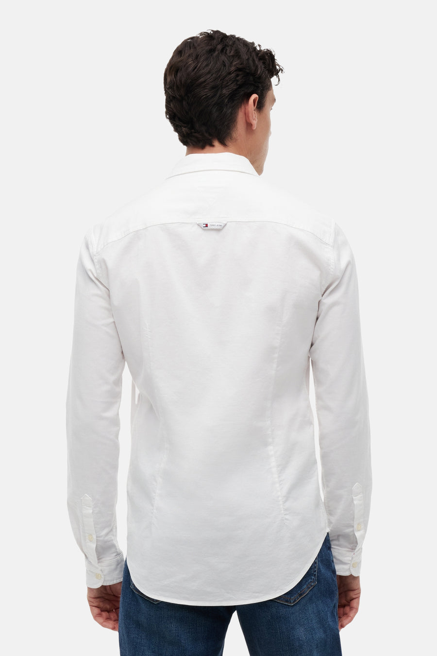 Wit hemd met lange mouwen van TOMMY JEANS, achterkant van een man, met klein TOMMY JEANS-logo op de rugpas en blauwe jeans.