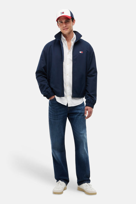 Chemise blanche à manches longues de TOMMY JEANS, portée sous une veste bleu marine, un jean et une casquette.
