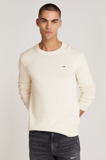 Beige trui met ronde hals van TOMMY JEANS, met subtiel logo op de borst, en gecombineerd met donkere jeans.
