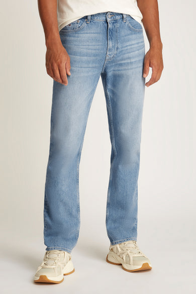 Jean slim de denim bleu clair de TOMMY JEANS, vu de l'arrière, avec un patch en cuir marron et des baskets blanches.