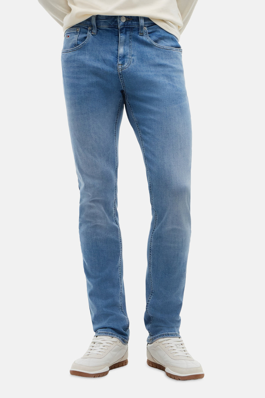 Jean slim bleu moyen de TOMMY JEANS, avec un léger délavage et une coupe ajustée, porté avec un t-shirt crème et des baskets blanches.