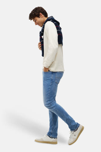 Jean slim en denim bleu moyen de TOMMY JEANS, porté par un mannequin avec un pull blanc et des baskets, profil latéral.