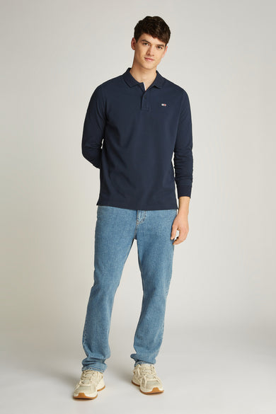 Blauwe polo met lange mouwen van TOMMY JEANS, gedragen met jeans en crèmekleurige sneakers.
