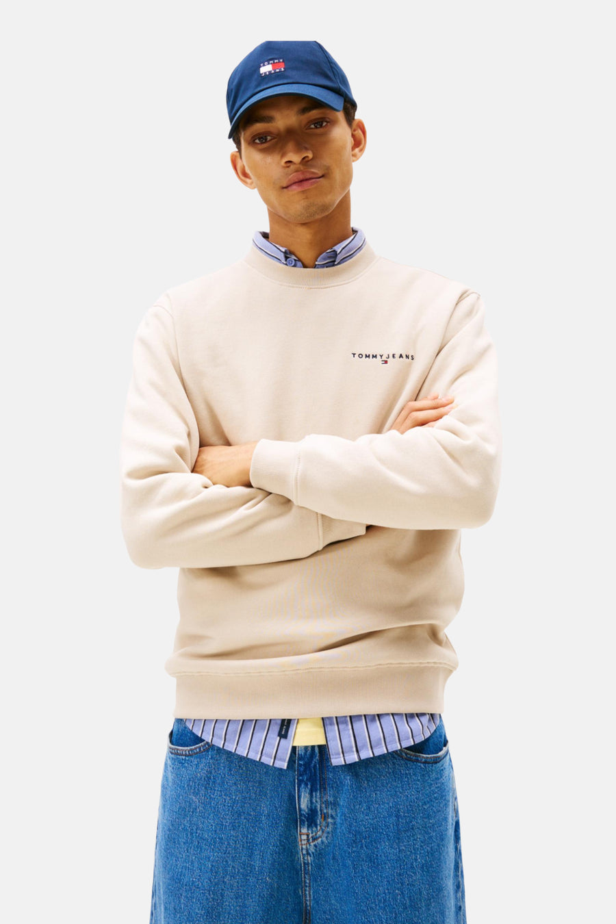 Een man draagt een blauwe pet, beige sweater en een gestreept overhemd, met gekruiste armen, en blauwe spijkerbroek.