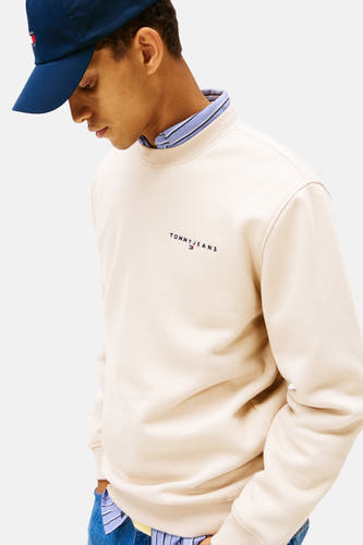 Gilet beige de TOMMY JEANS, avec un logo brodé, porté sur une chemise rayée et avec un chapeau bleu.