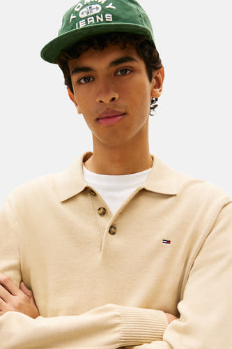 Beige trui van TOMMY JEANS, met kraag en logo op de borst, gedragen over een wit shirt en met een groene pet.
