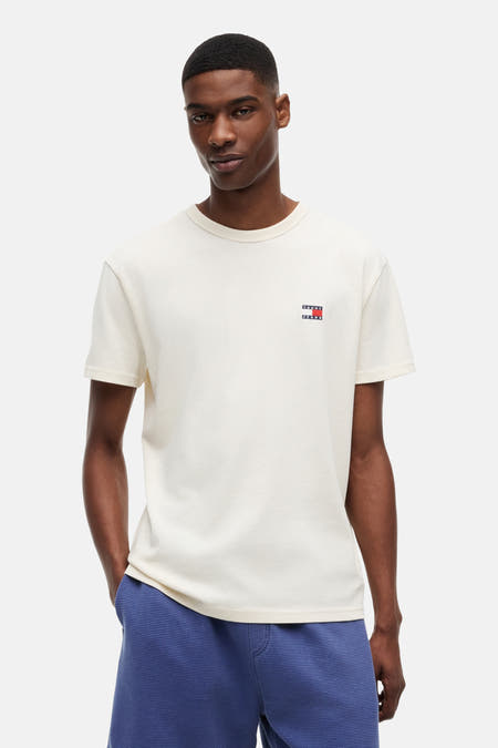 Crèmekleurig T-shirt met korte mouwen van TOMMY JEANS, met het kleine logovlaggetje links op de borst, gedragen door een man met blauwe wafelshorts.