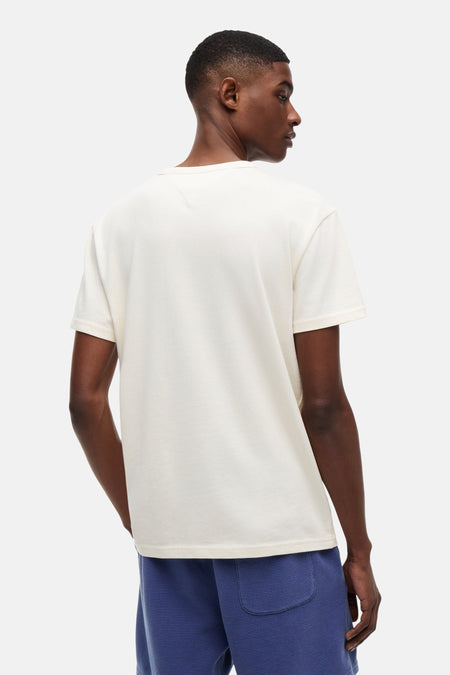 Crèmekleurig T-shirt met korte mouwen van TOMMY JEANS, achterkant zichtbaar met V-vormige stiksels in de nek, gedragen met blauwe wafelshorts.