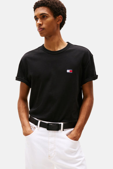 Zwart T-shirt met korte mouwen van TOMMY JEANS, met opgerolde mouwen en een klein logo op de borst, gedragen met een witte broek en zwarte riem.
