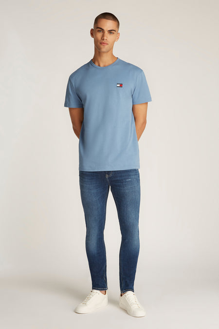 T-shirt bleu à manches courtes de TOMMY JEANS, avec un petit logo sur la poitrine, associé à des jeans foncés et des baskets blanches.
