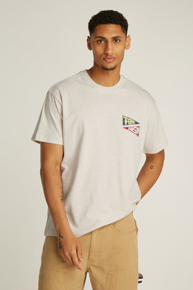 T-shirt écru à manches courtes de TOMMY JEANS, avec un petit logo drapeau sur la poitrine et le modèle porte un pantalon kaki.