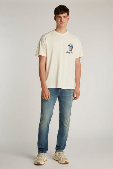 T-shirt blanc à manches courtes de Tommy Jeans, avec imprimé popcorn sur la poitrine, associé à des jeans bleus et des baskets crème.