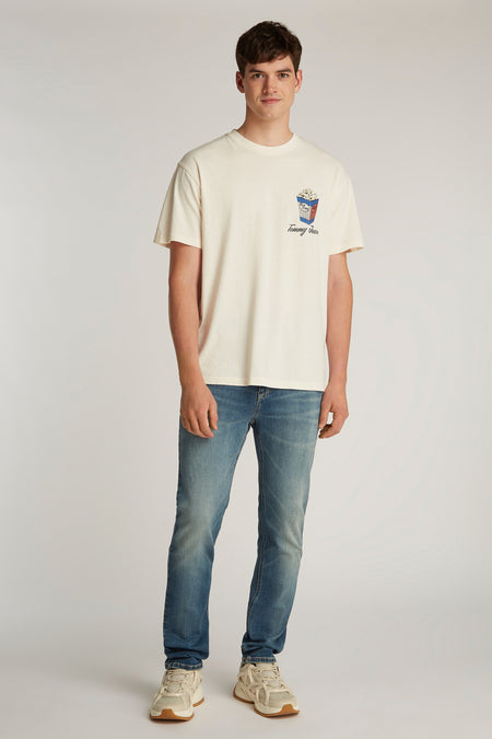 Wit T-shirt met korte mouwen van Tommy Jeans, met popcorn-print op de borst, gecombineerd met blauwe jeans en crèmekleurige sneakers.
