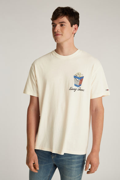 T-shirt blanc à manches courtes de Tommy Jeans, avec une illustration de popcorn sur la poitrine et porté avec des jeans bleus.