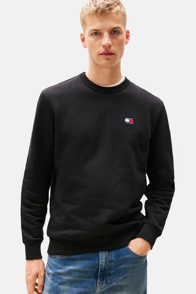 Pull-over noir de TOMMY JEANS, avec un petit logo sur la poitrine, porté avec une chemise blanche et des jeans.