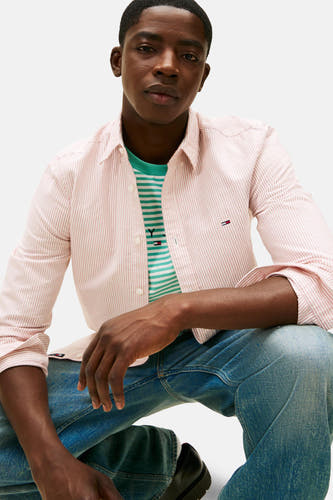 Rood gestreept overhemd met lange mouwen van TOMMY JEANS, met een geborduurd logo, gedragen over een gestreepte top en jeans.
