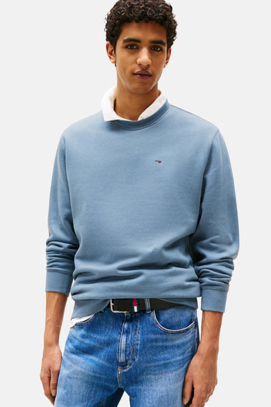 Blauwe sweater van TOMMY JEANS, met lange mouwen en een klein logo op de borst, gedragen over een wit overhemd en met een jeans.
