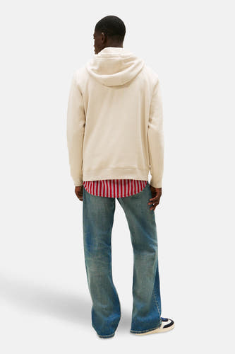 Beige sweater met kap van TOMMY JEANS, met een rood-wit gestreept overhemd eronder en blauwe jeans, gezien van achteren.
